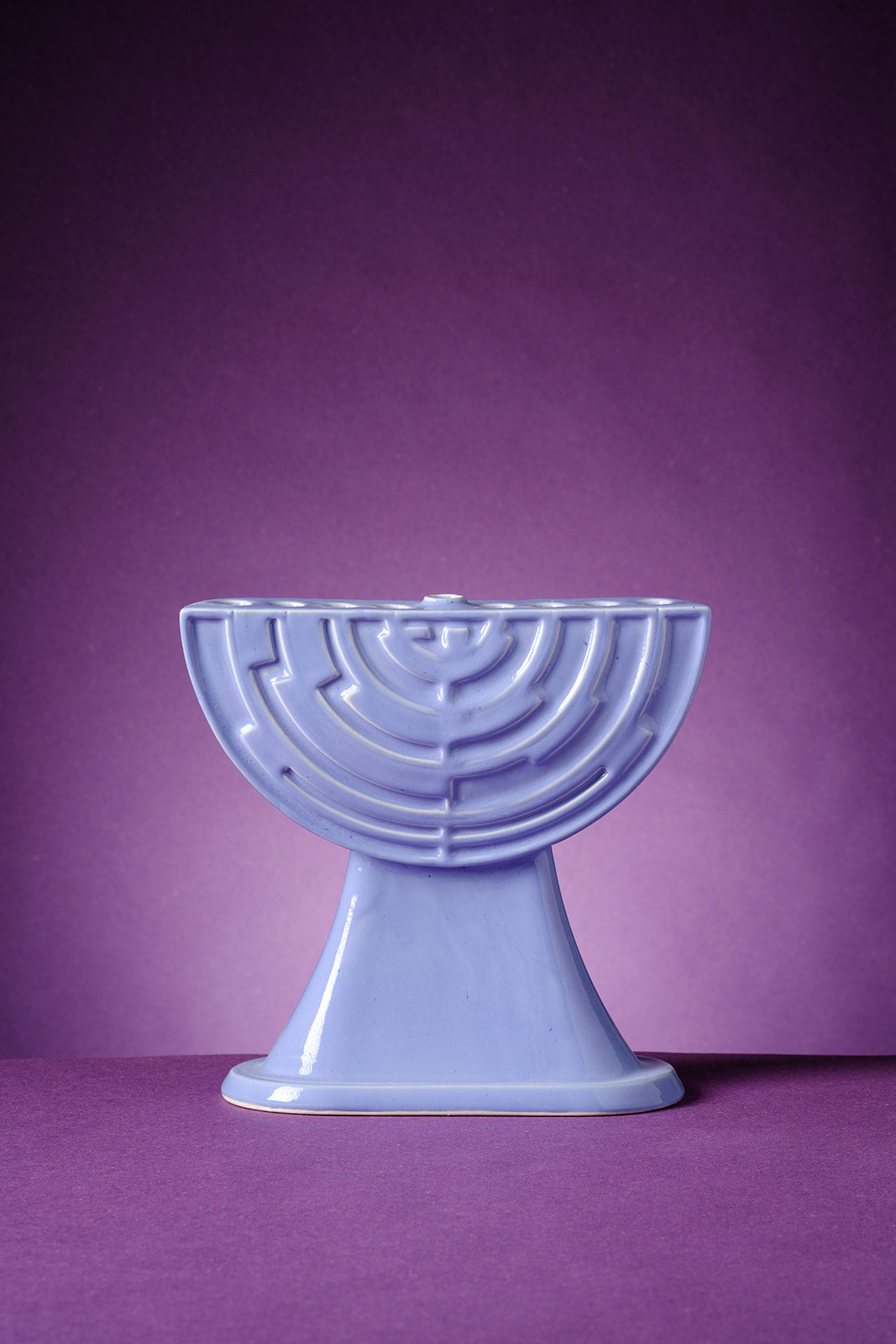 A sculptural lavender ceramic menorah by ABS Objects, handmade in Tel Aviv. The menorah features a semi-circular form with concentric geometric reliefs inspired by the logo of Israel’s first television channel. Its glossy surface and modernist silhouette reflect the studio's fusion of contemporary design, local history, and Hebrew visual culture. Photographed against a deep purple background, the piece embodies a modern take on Judaica with cultural and aesthetic depth.