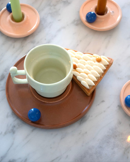 Green mug and pastry on a brown plate with candle holders and blue candle holders on a marble surface