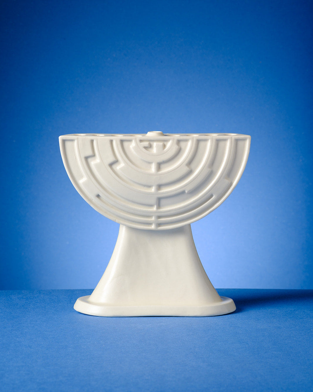 A modern ceramic menorah with a geometric, maze-like pattern stands on a green surface against a warm yellow-orange gradient background. The menorah holds nine lit red candles, with soft glowing flames.









