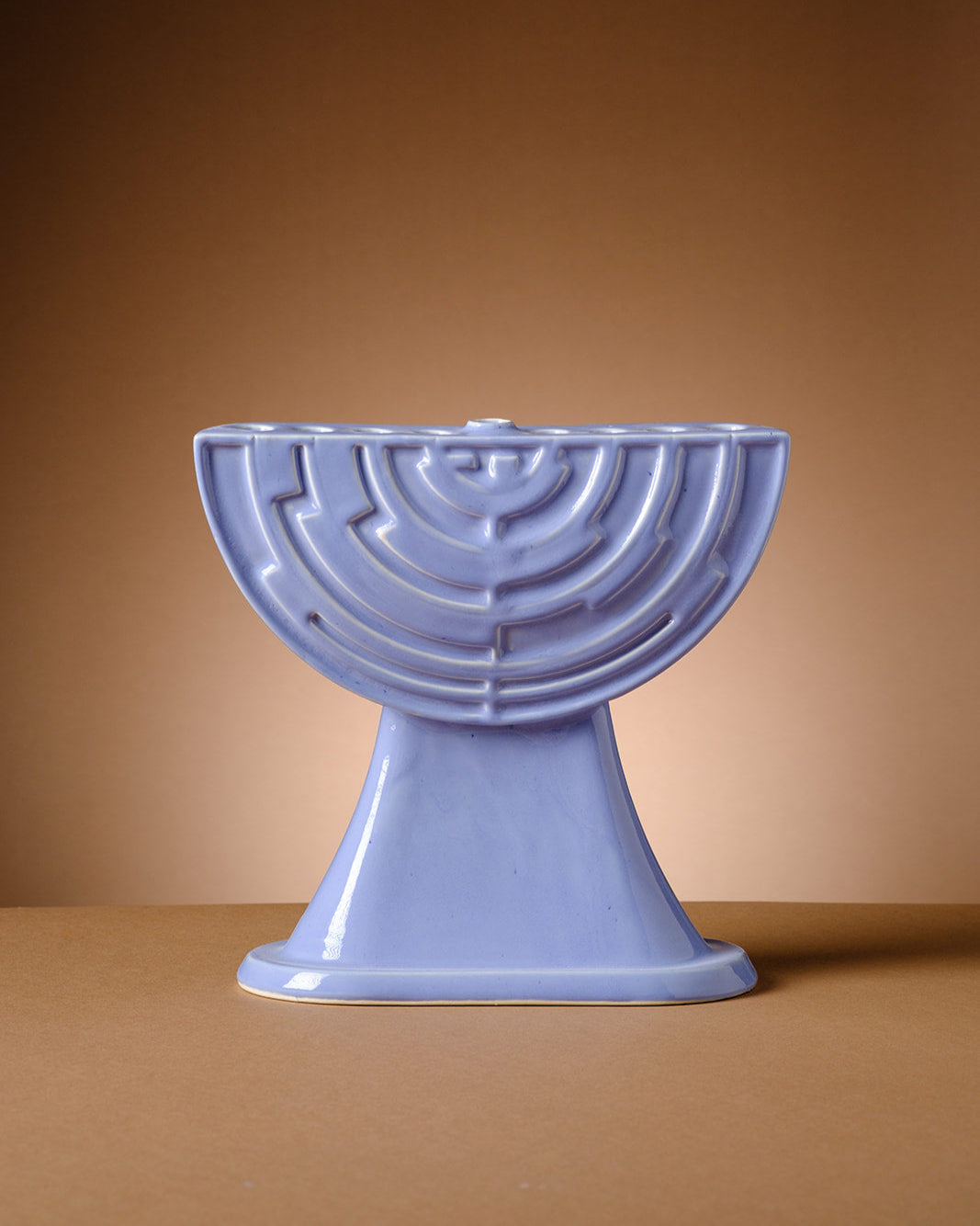 A sculptural lavender ceramic menorah by ABS Objects, handmade in Tel Aviv. The menorah features a semi-circular form with concentric geometric reliefs inspired by the logo of Israel’s first television channel. Its glossy surface and modernist silhouette reflect the studio's fusion of contemporary design, local history, and Hebrew visual culture. Photographed against a deep purple background, the piece embodies a modern take on Judaica with cultural and aesthetic depth.