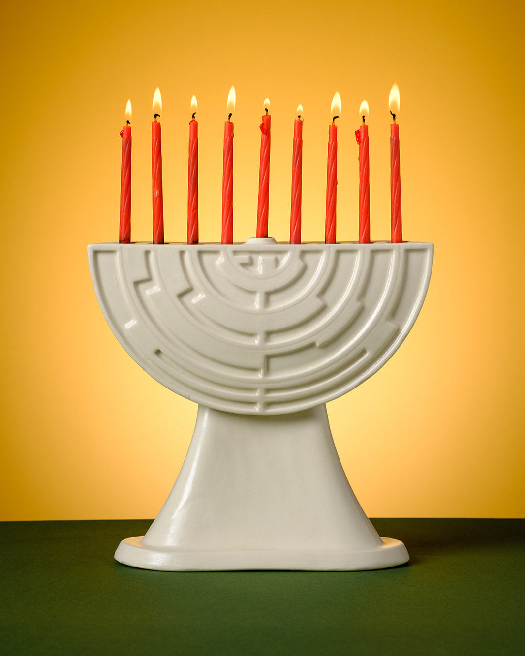 A modern ceramic menorah with a geometric, maze-like pattern stands on a green surface against a warm yellow-orange gradient background. The menorah holds nine lit red candles, with soft glowing flames.